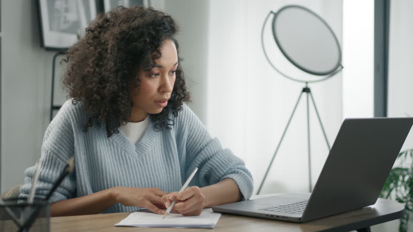 Focused and concentrated african female student e learning online on internet website. Young woman of color working remotely at home office desk, makes notes in work document, looks at laptop computer | Shutterstock HD Video #1102675493