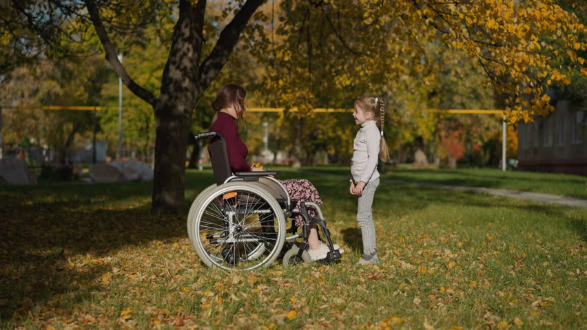 Young mother with legs injury throws bunch of dry leaves up above daughter. Preschooler girl raises hands rejoicing at game in autumn park side view