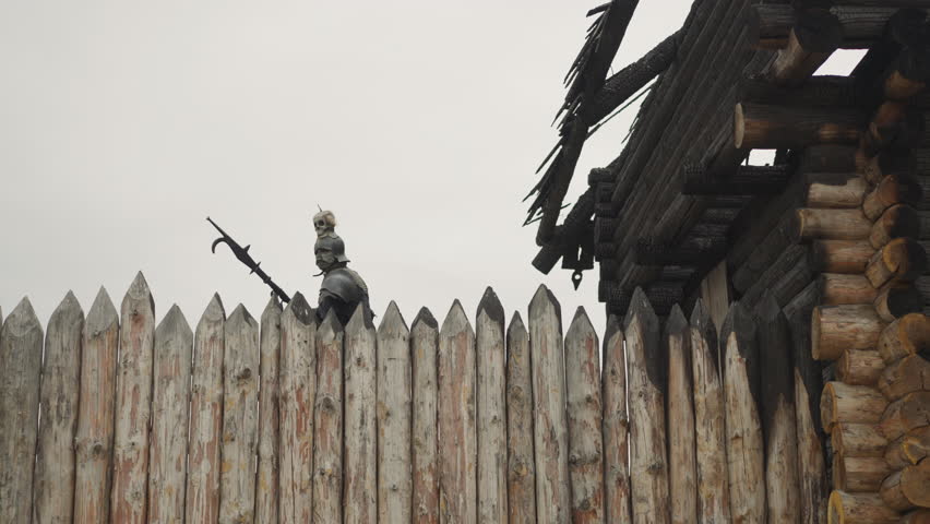 Evil knight with spear walks near burnt wooden house behind fence rain. Medieval demonic man looks for people in ancient village under grey sky low angle shot
