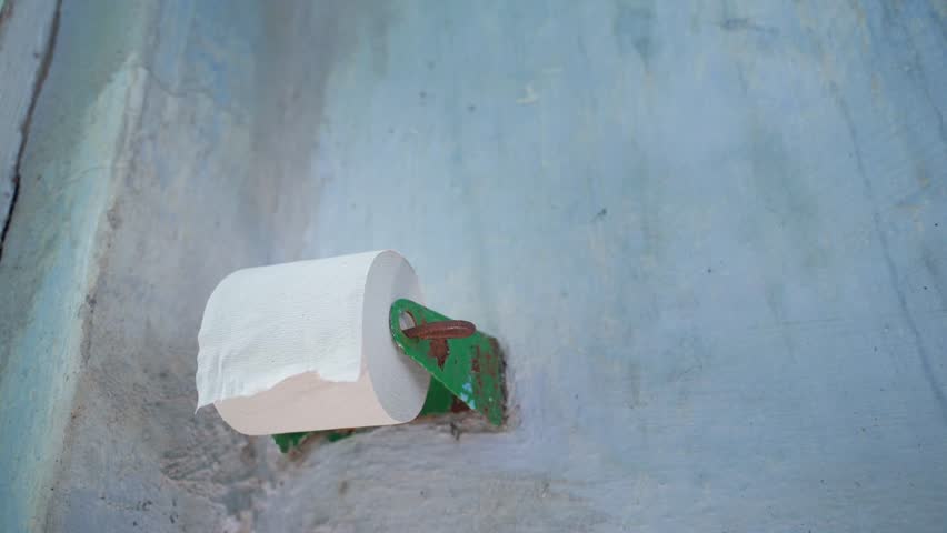 A hand reaches for a toilet paper holder in a rural outdoor toilet. A roll of single-layer toilet paper is being pulled, close-up