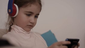 Little Girl With Headphones Playing Video Game on Mobile Phone. Teenager Plays Video Game On Smartphone. Child Happy Playing Games With Phone. Harm for Mental Kid Health Eyesight Gambling Addiction