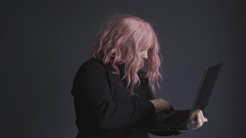 A man in a pink wig stands and works at a computer, typing. Side view