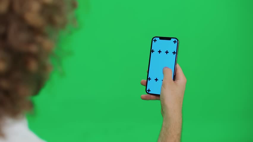 Back view, young man uses smartphone, scrolls through the images and swipe across the blue screen, green background, vertical view.