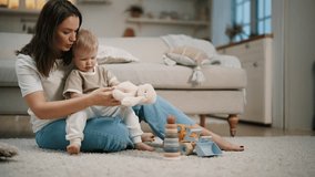 Emotional positive mom and toddler daughter plays together sits on floor in living room with teddy hare. Smiling mother holds baby girl on knees shows toys parts. Motherhood, maternity leave concept. - Powered by Shutterstock - Get 15% off with code: PIKWIZARD15