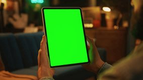 In the evening hands man holding and tapping on a tablet computer with a vertical green screen. Mock up. Sofa on background