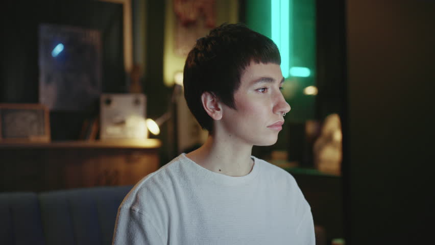 Portrait of smart confident young Caucasian woman with piercing sitting at computer and working indoors. Businesswoman looking at camera in cozy environment of loft office with neon light