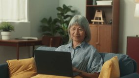 Happy attractive Asian elderly woman using laptop notebook computer video conference call in cozy living room. Happy old aged female using technology chatting talking with family or friends at home