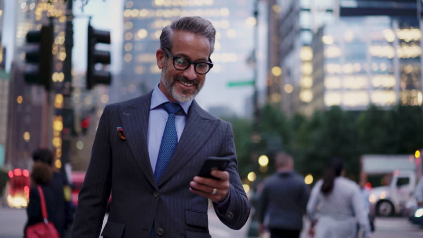 Handsome and Successful business man in stylish suit looking  at smartphone screen while walks outdoors in city. Prosperous smiling entrepreneur in eye glasses feels confident and prosperous 