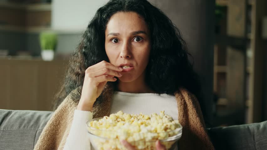 Camera view of addicted focused young hispanic woman watching interesting TV program film movie and eating popcorn alone at home Enchanted curly girl cannot take her eyes off the screen indoors Royalty-Free Stock Footage #1102716083