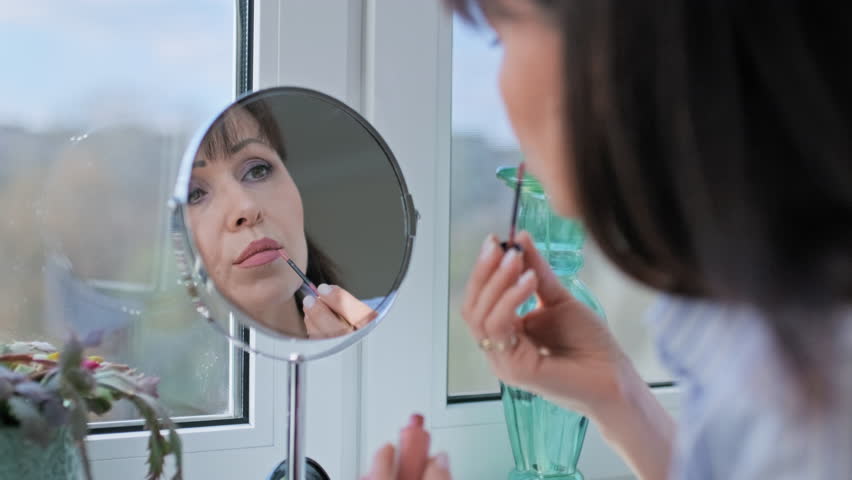 Middle aged woman doing makeup, paints lips with long-lasting lipstick looking in mirror