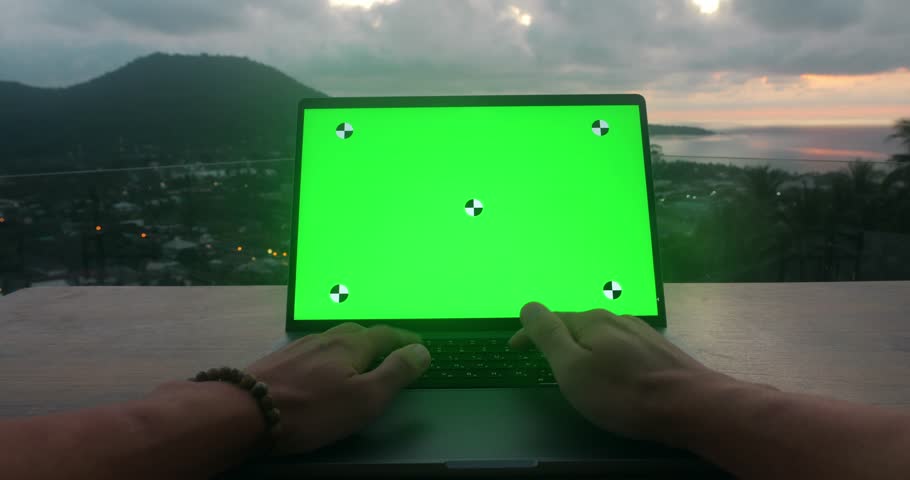 Male hands typing on laptop with green mockup screen at outdoors workplace at sunset on vacations. Business man using laptop computer with green screen, on the roof at sea view. POV shot.