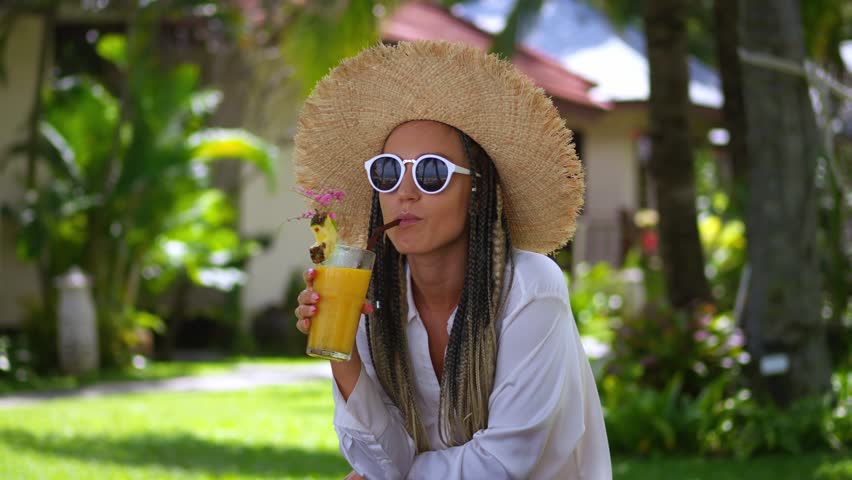 Portrait glad woman tourist enjoying summer cold drink banana fresh juice, resting on summer vacation. Woman in sunglasses and hat drinking orange juice at summertime. Concept resting on vacation