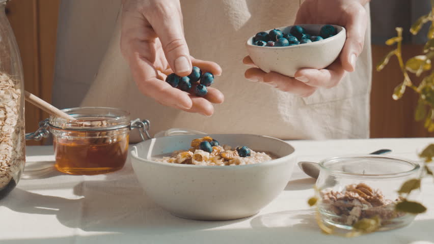 Woman preparing healthy dieting vegan nutritious breakfast. Female hand putting blueberries in the bowl with oatmeal porridge with walnuts and honey. Royalty-Free Stock Footage #1102738107