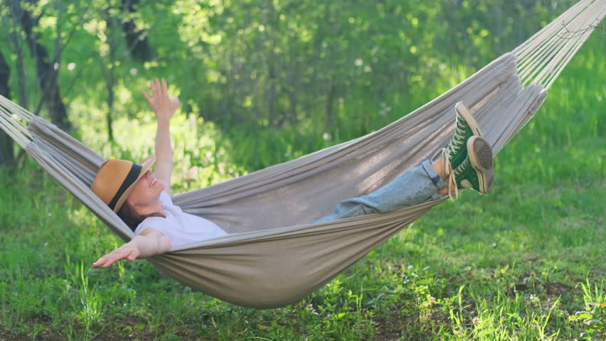 Cheerful laughing woman having fun swinging in a hammock in sunshine. Young girl in a great mood enjoying her summer vacation in a green garden. Happy emotions, people joy concept. Royalty-Free Stock Footage #1102740083