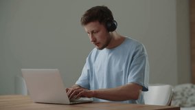 A young man in a blue T-shirt is typing on a laptop keyboard while sitting at a table. A male freelancer works remotely on a laptop and listens to music on headphones.