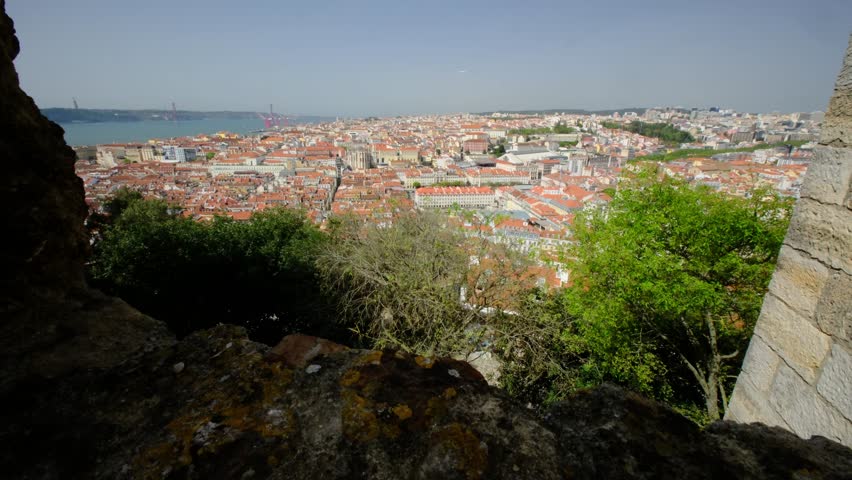 An extreme wide angle view of a modern city, with beautiful red tiled roofs, as seen through an old castle walls. a distant bridge and a river 
are also visible. Lisbon, Portugal.
