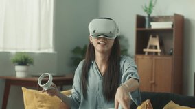 Young Beautiful Asian woman putting on Virtual Reality Headset technology using Joysticks enjoy focus on playing VR Battle Game on sofa at cozy living room in the holiday. Lifestyle at home concept