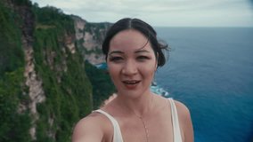 A cute travel blogger Asian women hold camera in arms and shooting selfie video shows audience place on edge of cliff above oceans. Travel trip for female broadcasting on social network. Emotions