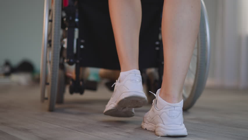 Legs of young woman pushing wheelchair with patient with disability on blurred background. Nurse takes care of person with paralysis at home closeup Royalty-Free Stock Footage #1102755167
