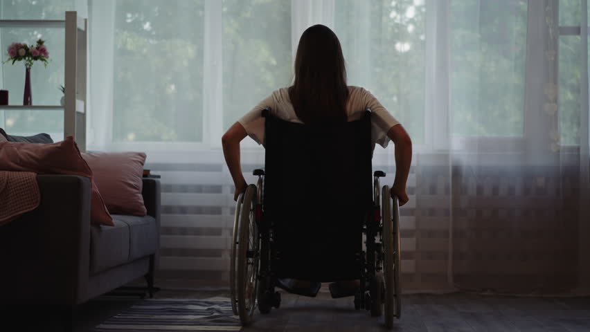 Lonely woman with disability moves wheelchair to look out bright window. Depressed brunette lady spends time home after car accident backside view Royalty-Free Stock Footage #1102755191