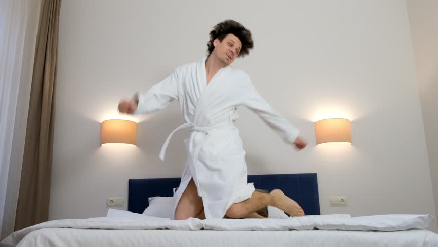 Happy young brunet man in white bathrobe is smiling jumping and falling in bed in hotel room in slow motion. He is resting on his vacations, weekend. Comfort leaving in journey concept.