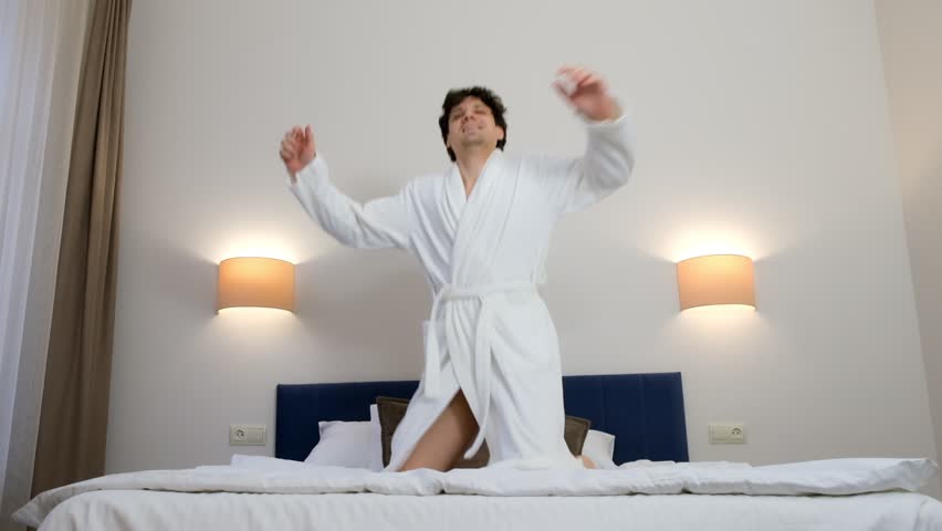 Happy young brunet man in white bathrobe is smiling and jumping in bed in hotel room in slow motion. He is resting on his vacations, weekend. Comfort leaving in journey concept.