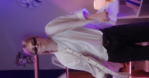 Vertical video of man recording dance video on phone camera in living room. Young male teenager dancing at home for musical clip. Blogger making music content for channel, wearing sunglasses.