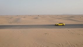 A yellow car is driving along an asphalt road in the desert, raising sand dust behind it. Drone Video