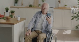 Emotional grandfather making online video call with smartphone speaking and gesturing sitting in wheelchair in kitchen at home. Senior people and modern technology concept.