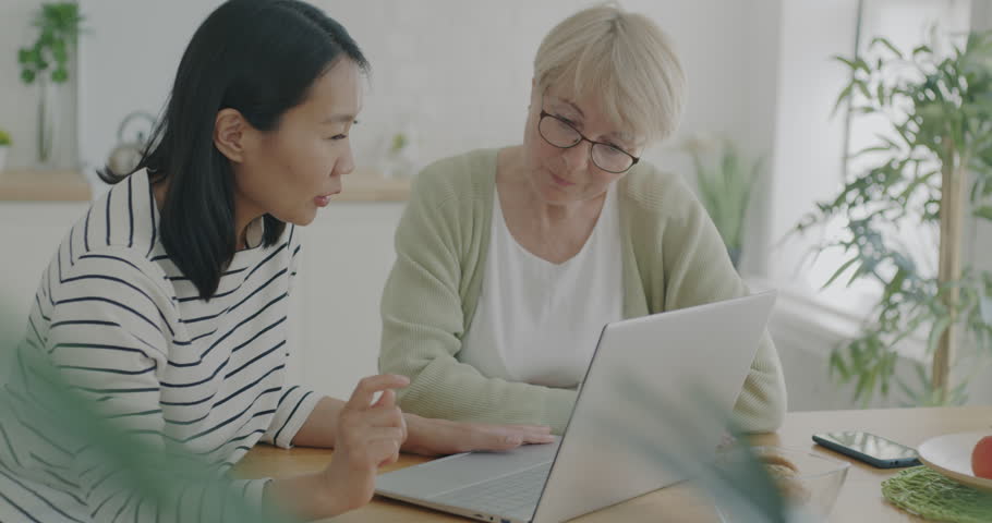 Female volunteer helping senior woman with laptop teaching technology talking and using computer at home in kitchen. Lifestyle and volunteering service concpt.