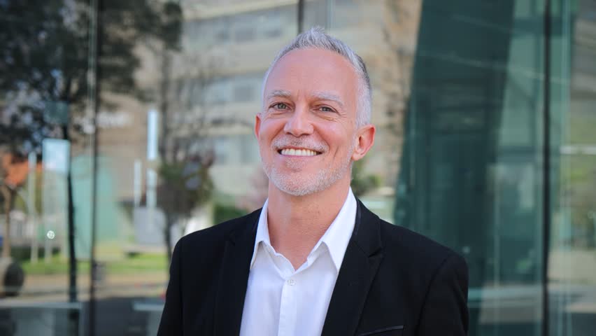 Close up portrait of mature adult business man with gray hair and suit smiling and looking at camera with succesful attitude. Happy corporate lawyer with white perfect teeth standing at workspace