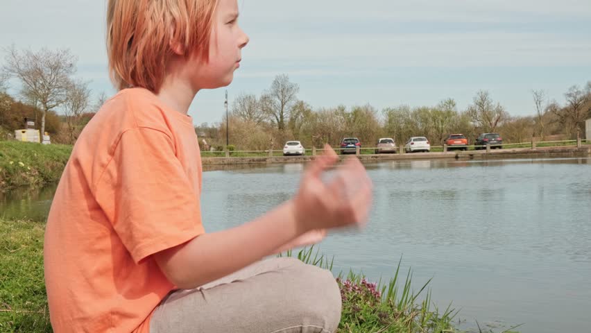 A young boy is meditating outdoors. A boy with red hair sits in a lotus position near a lake. Relaxation, rejuvenation. Side view.