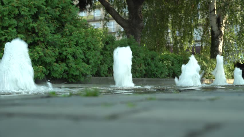 Pedestrian fountain cascading in the city in sunny weather. Movement transparent splash in green foliage background. Sparkling gush water of liquid drops detail. Refreshing cool on hot day close-up.