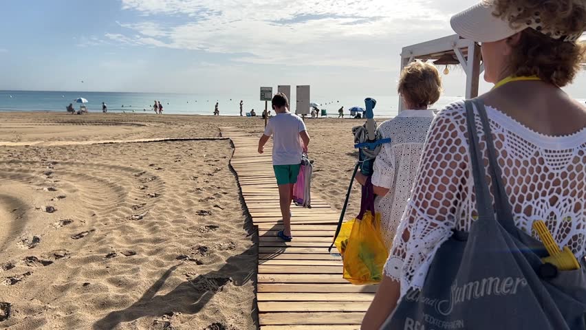 Slow motion. Two women, one adult and one older, and a teenager walk with beach chairs along a wooden walkway. Summer vacation concept