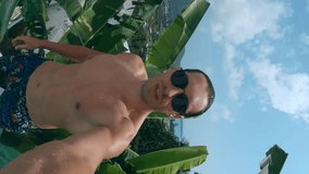 Vertical selfie video man filming himself on action camera jumping in pool during summer days. Active man on glasses jumps in pool and filming himself as he dives underwater during summer days