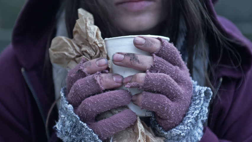 Homeless young woman, shivering from the cold, holds a paper cup of some kind of drink in her dirty hands.