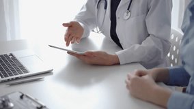 Doctor and patient sitting at the white desk near flair window in clinic. Unknown female physician wearing a white coat uses tablet computer for filling up medical record form. Medicine concept