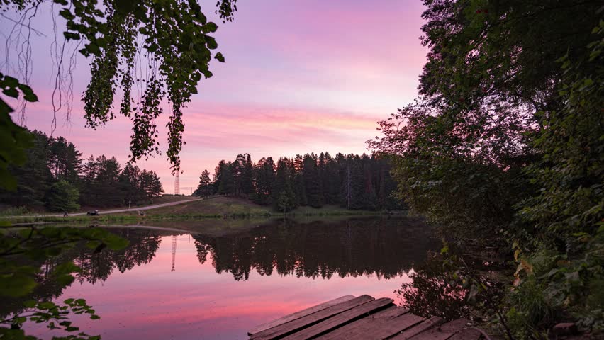Time-lapse sunrise over beautiful lake. Water surface in fast motion. Warm sky with rolling clouds. Trees silhouette woodland on horizon. Early morning shot in timelapse. Bright sun rises over skyline