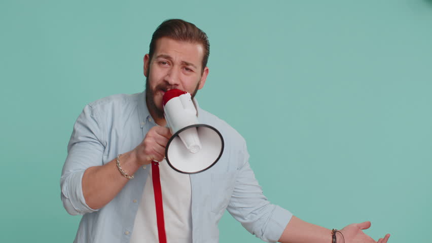Lebanese man talking with megaphone, proclaiming news, loudly announcing advertisement, warning using loudspeaker to shout speech, pointing empty place. Copy-space. Handsome guy on blue background