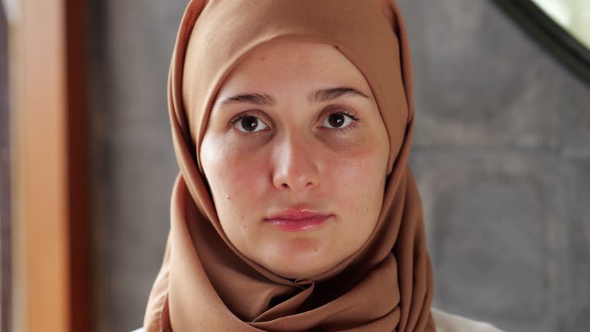 Young woman in hijab covering her hair looks directly into the camera, close portrait shot. Lady smiling, and we can see the braces on her teeth. She looks slightly shy and show natural emotion