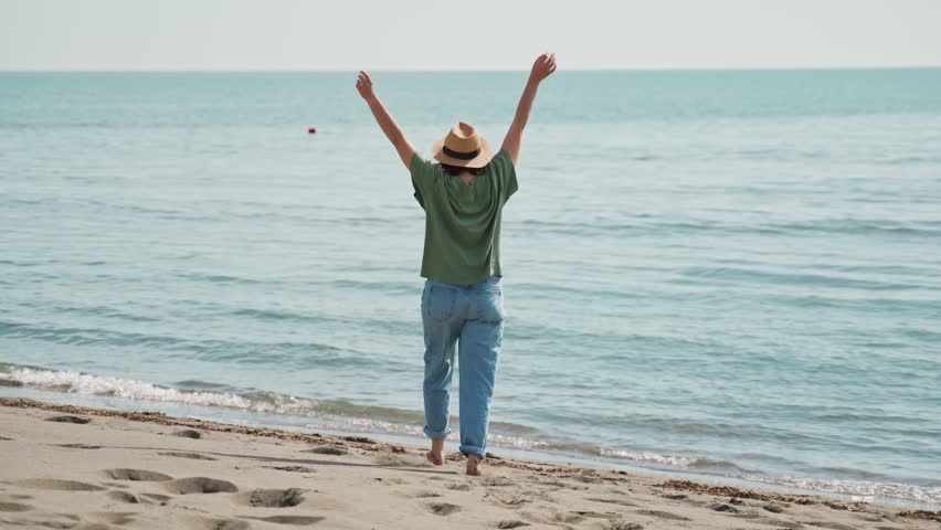 An adult Caucasian woman in a hat walking along a sandy beach toward the sea on a sunny day.