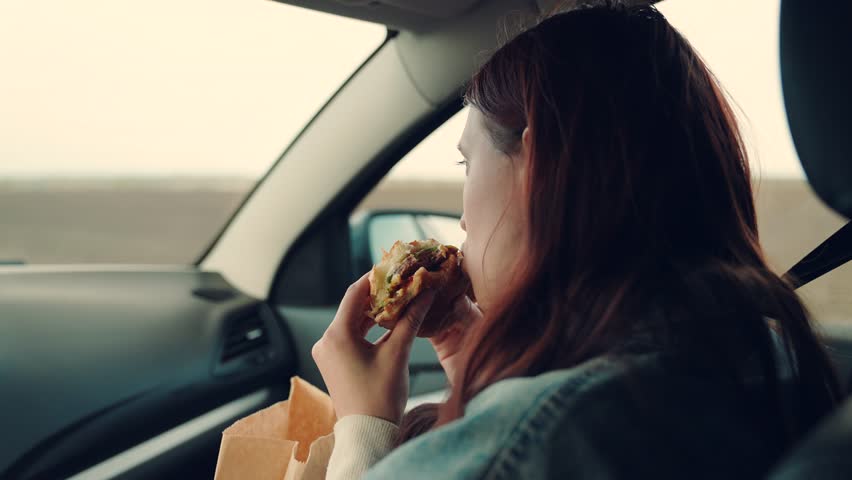 Girl passenger eats burger in car. Delicious food bun with cutlet. Young hungry woman eats roll with meatball while sitting in car. Offspring travels, having snack on road without leaving car.Tourist Royalty-Free Stock Footage #1102797005