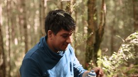 A young latino man drinking water from a bottle in the forest. Small break during hiking in the mountains. Healthy lifestyle and activity to enjoy adventures in nature concept in 4K