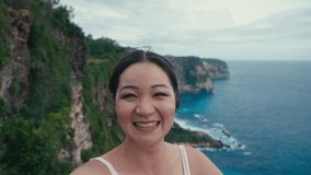 A cute travel blogger Asian women hold camera in arms and shooting selfie video shows audience place on edge of cliff above oceans. Travel trip for female broadcasting online on social network.