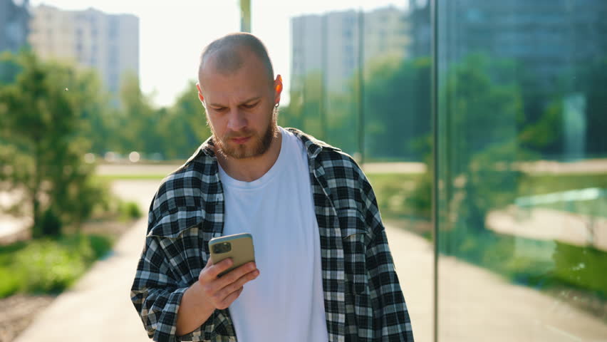 Handsome Caucasian Man Using Smartphone While Walking Near Office Center. Man Swiping Apps, Texting with Friends Outside. Businessman Working on the Smartphone, Communicating with Clients