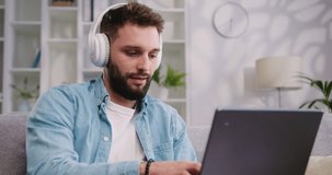 Good-looking confident modern smart man in headphones sitting on the couch at home and working on laptop. Male freelancer or student listens to an online conference at home.