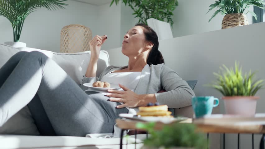 Video of beautiful happy pregnant woman in panties eating chocolate truffles lying on sofa at home Royalty-Free Stock Footage #1102812513