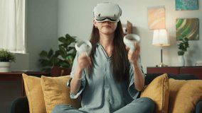 Young Beautiful Asian woman putting on Virtual Reality Headset enjoy using Joysticks focus on playing VR game technology feeling excited and fun on sofa at cozy home living room in the holiday