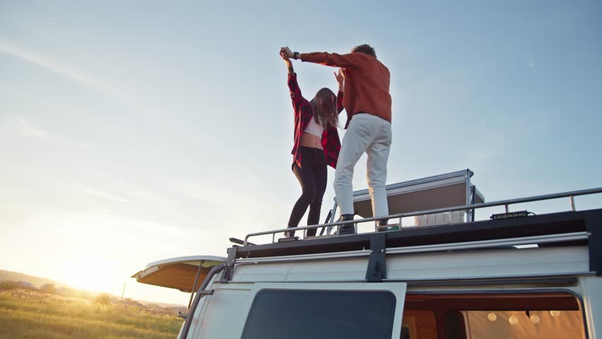 Beautiful caucasian pair dancing on their camper van rooftop. Young girl and boy enjoying their romantic road trip. Countryside background, summer evening, relaxing on fresh air Royalty-Free Stock Footage #1102815877