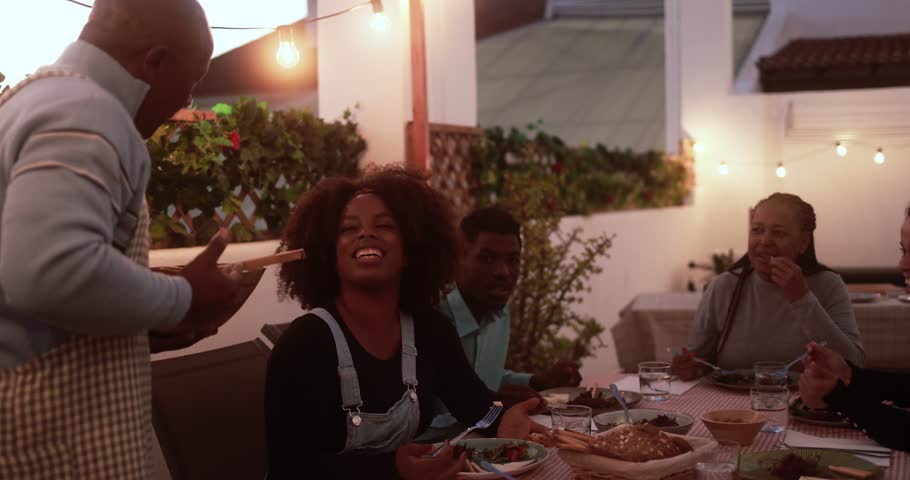 Happy african family doing dinner while eating healthy food at home patio - Father serves food - Lifestyle home moments and summer concept Royalty-Free Stock Footage #1102816765